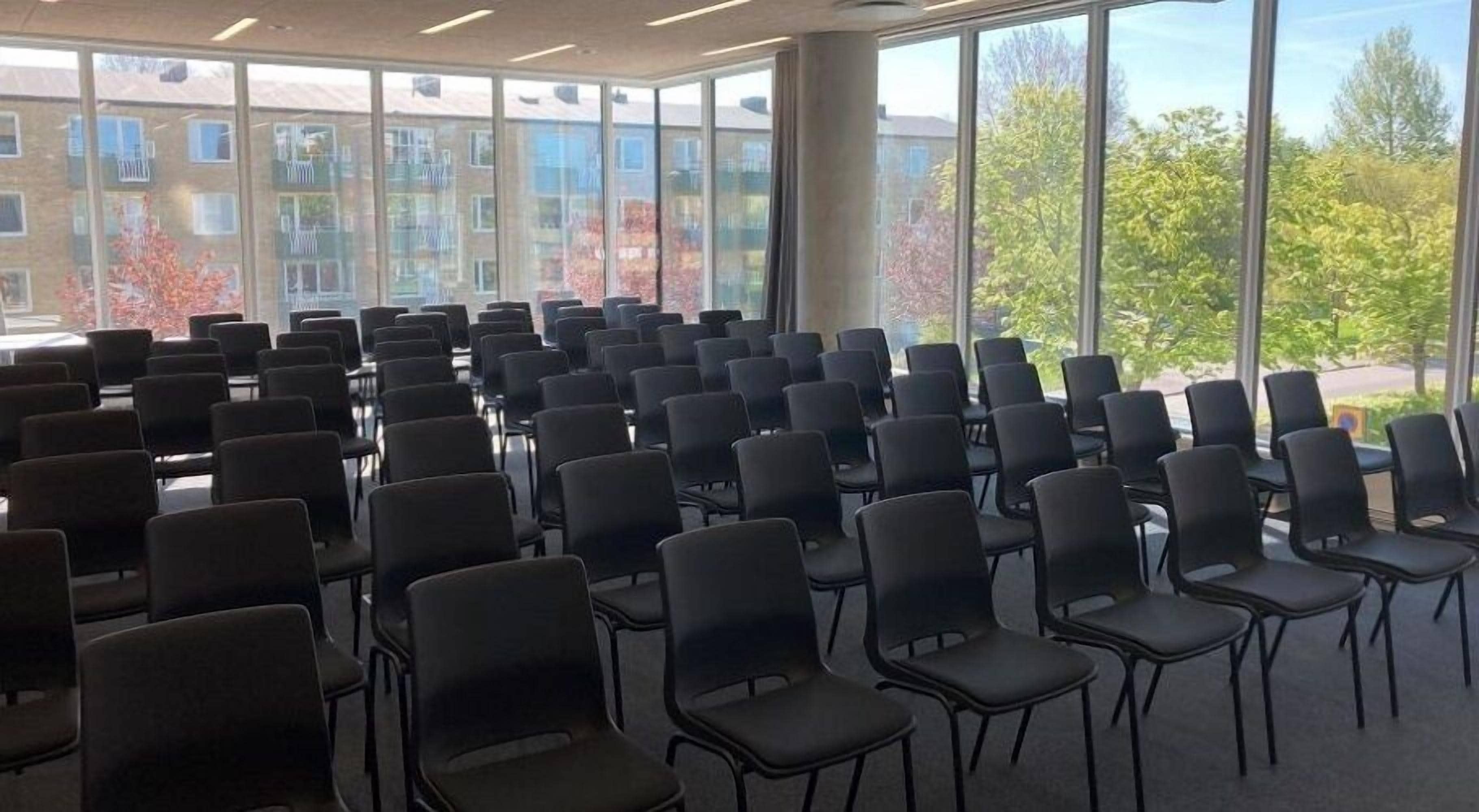 A conference room with rows of chairs, large windows with a view of buildings and trees, and a high ceiling.
