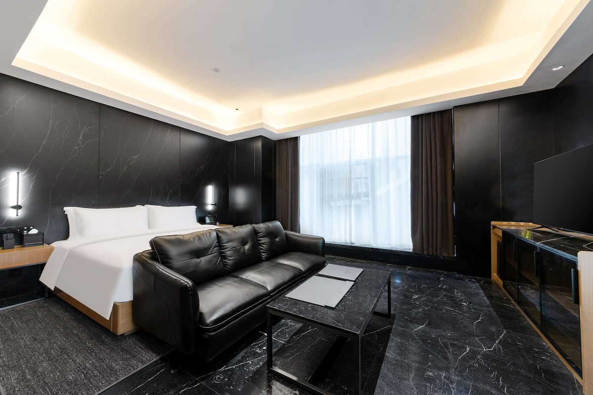 Habitación de hotel moderna con sofá de cuero negro, cama con ropa blanca, piso de baldosas de mármol, una mesita de centro de vidrio y un ventanal con cortinas.