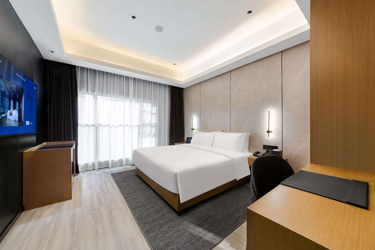 Habitación de hotel moderna con una cama grande, un escritorio, un televisor y un ventanal con cortinas.
