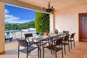 A dining area with a table set for six, surrounded by wicker chairs, a chandelier, and a view of a green wall and landscape.