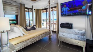 A bedroom with a large bed, a TV mounted on the wall, and a view of the city through the windows.