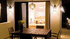 A dining area with a wooden table and chairs, a vase with flowers, and a view into a room with a bed and curtains.