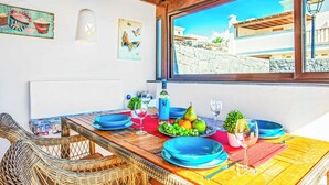 Un coin repas avec une table en bois dressée pour deux personnes, agrémentée d’assiettes bleues, d’une bouteille de vin et d’un panier de fruits.