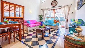 Un salon avec un sol à carreaux, des meubles en bois, un canapé agrémenté de coussins colorés, une table à manger avec des chaises, une table basse, une horloge au mur et une fenêtre avec des rideaux.