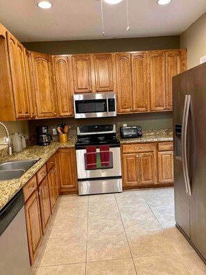 A kitchen with wooden cabinets, stainless steel appliances including a microwave, oven, and refrigerator, a granite countertop, and a tiled floor.
