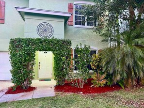 A building with a circular decorative element above the entrance, a door, and a well-maintained garden with various plants and a palm tree.