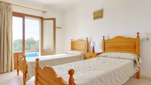 A bedroom with two wooden beds, a window with curtains, a bedside table with a lamp, and a view of a swimming pool. - Villa La Pinta - Five Bedroom Villa, Sleeps 10 (Cala d'Or)
