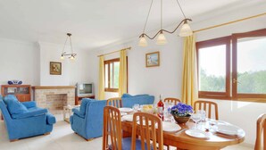 A dining area with a wooden table set for four, blue upholstered chairs, and a fireplace in the background. - Villa La Pinta - Five Bedroom Villa, Sleeps 10 (Cala d'Or)