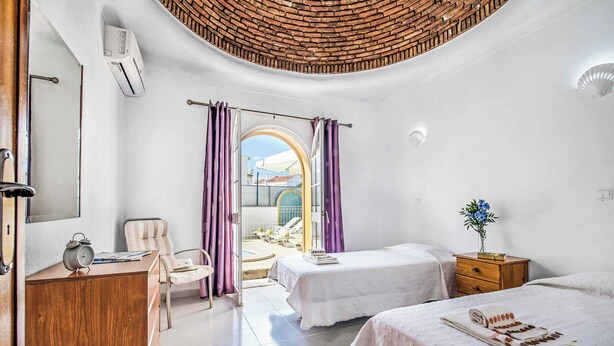 Ein Hotelzimmer mit zwei Betten, einem Schreibtisch, einem Stuhl und Blick auf den Poolbereich durch eine offene Tür.