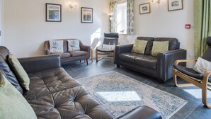 A living room with a brown leather sofa, a brown leather armchair, and a wooden chair with a cushion. There are two framed pictures on the wall and a lamp on the side table.