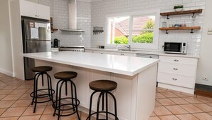 Een moderne keuken met witte kasten, een groot eiland met krukken, een roestvrijstalen koelkast, een magnetron en een raam met uitzicht op groen.