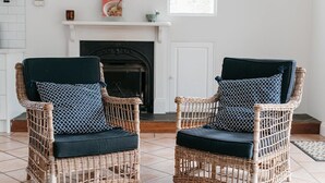 Twee rieten stoelen met donkere kussens in een kamer met een witte schoorsteen en een betegelde vloer.
