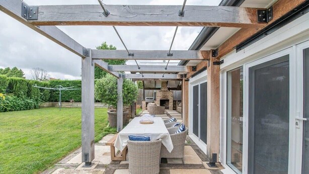 Een terras buiten met een tafel en stoelen, een pergola en uitzicht op een met baksteen beklede open haard.