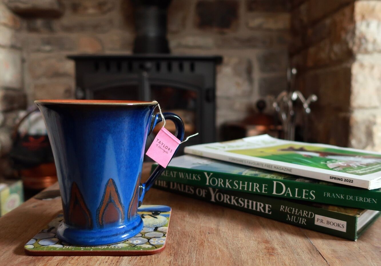 A blue ceramic cup with a tag on it, placed on a coaster, next to a stack of books titled 'Yorkshire Dales' by Frank Duerden and 'The Dales of Yorkshire' by Richard Muir, on a wooden table with a fireplace in the background.