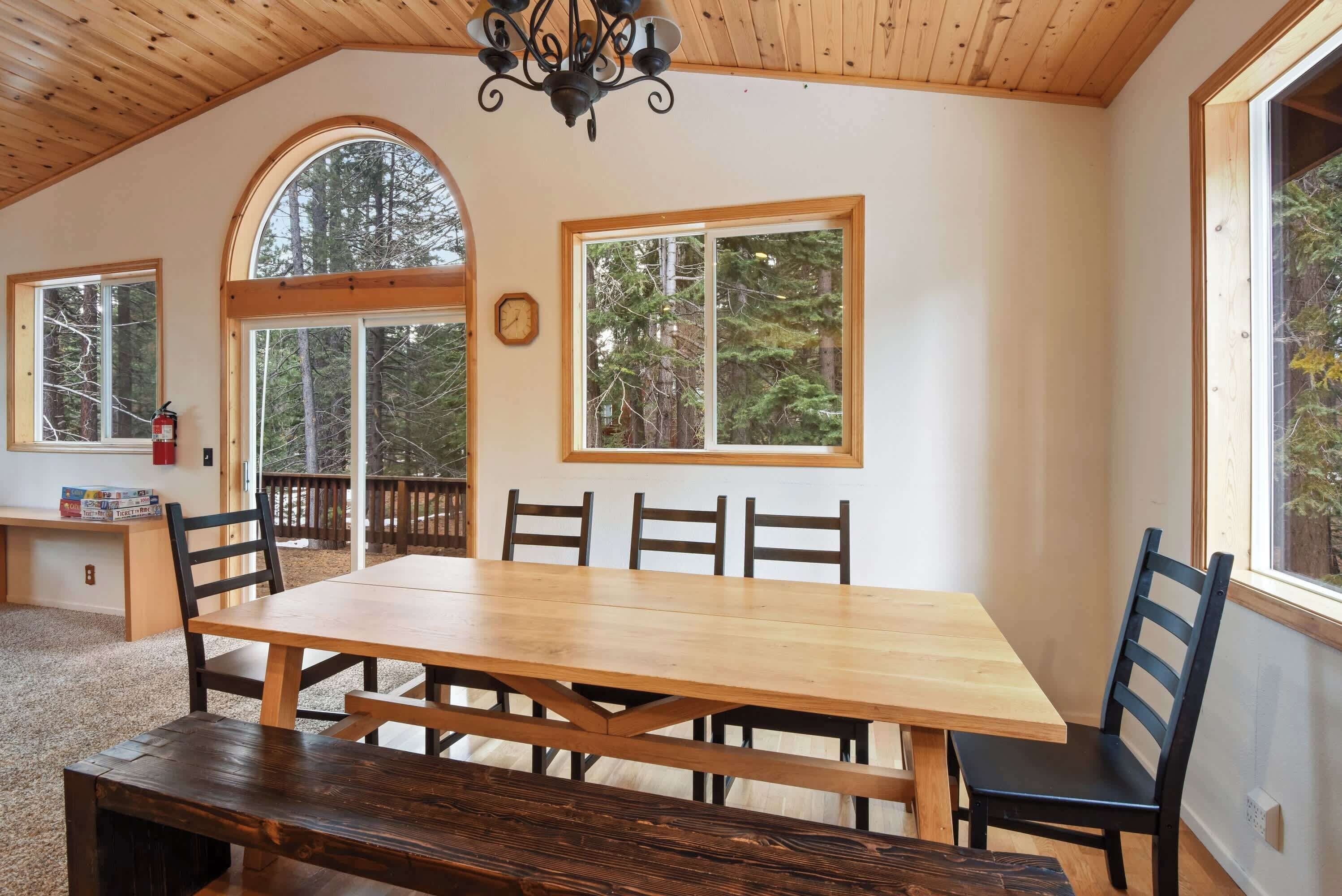 Un comedor con mesa y sillas de madera, ventanas amplias con vista a árboles y un reloj en la pared.