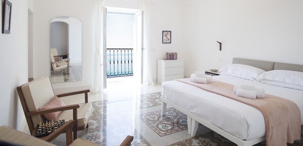A bright, minimalist bedroom with a large bed, a chair, a nightstand, and a view of the outside through a window.