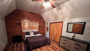 Ein Schlafzimmer mit Holzboden, einem Bett mit dunkler Bettdecke, einer Kommode mit Schubladen, einer Gitarre an der Wand und einer Tür mit Spiegel.