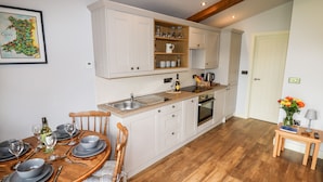 A compact kitchen with white cabinets, a wooden dining table set for four, and a small side table with a vase of flowers. - Celyn (Ruthin)