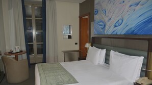 Habitación de hotel con cama, escritorio, silla, un cuadro y puerta que da a un balcón.