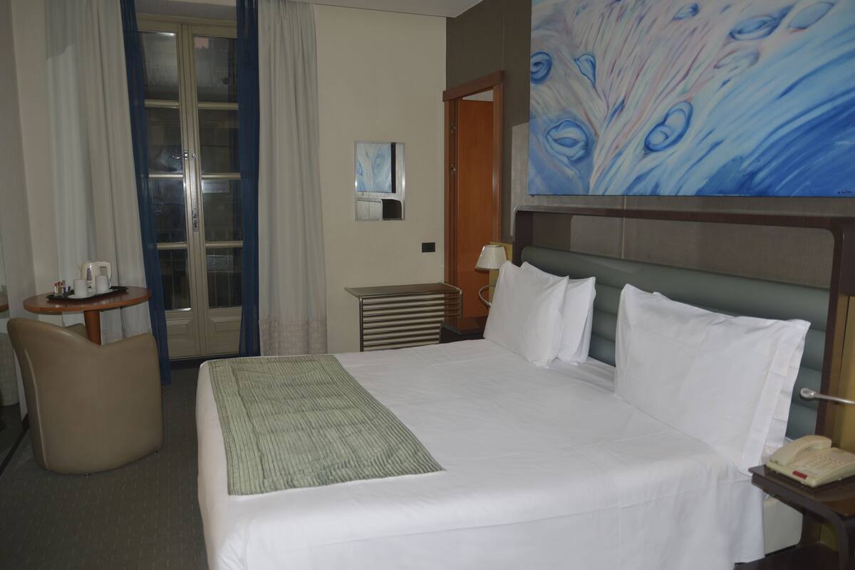 Habitación de hotel con cama, escritorio, silla, un cuadro y puerta que da a un balcón.