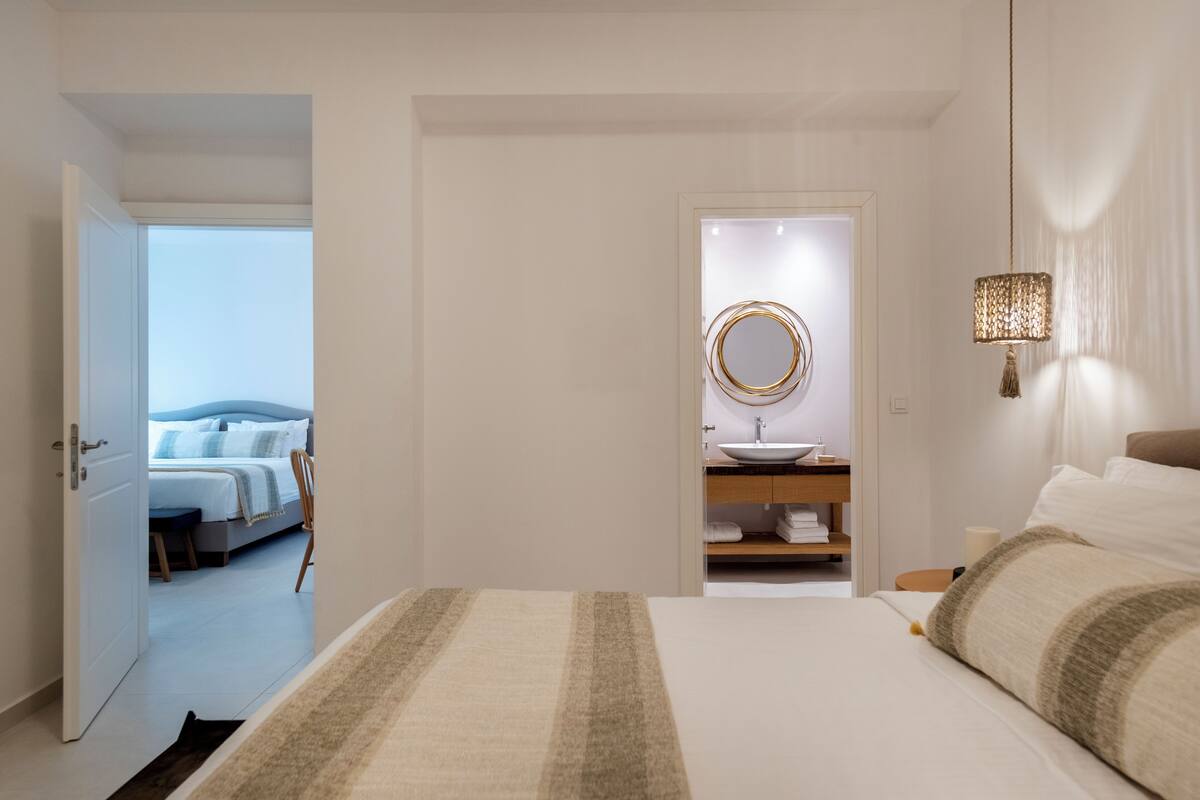 Ein Hotelzimmer mit Bett, Nachttisch, Lampe, Badezimmer mit Waschbecken und Spiegel sowie Blick auf das angrenzende Zimmer.