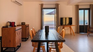 A dining area with a wooden table, chairs, a TV, and a view of the sea through a window. - La Maltese Villas Santorini (Santorini)