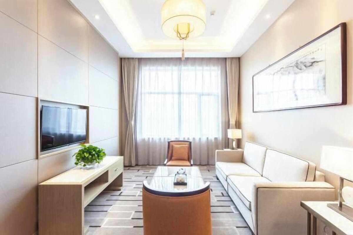 A modern hotel room with a flat-screen TV, a sofa, a coffee table, a TV stand with a plant, and a large window with curtains.