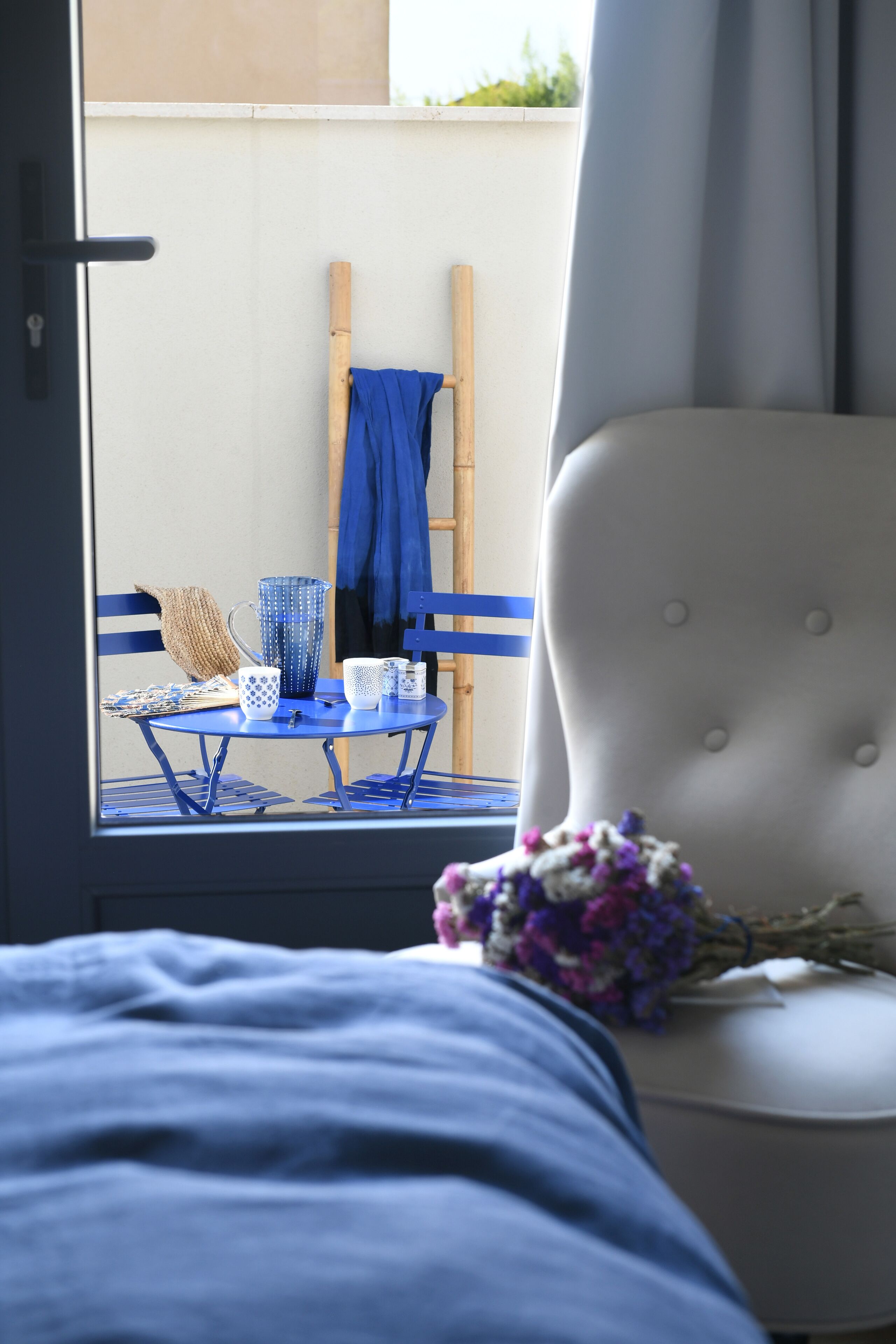A room with a bed, a chair, a table with a blue tablecloth, a blue chair, a white chair, a blue vase, and a wooden ladder leaning against a wall.