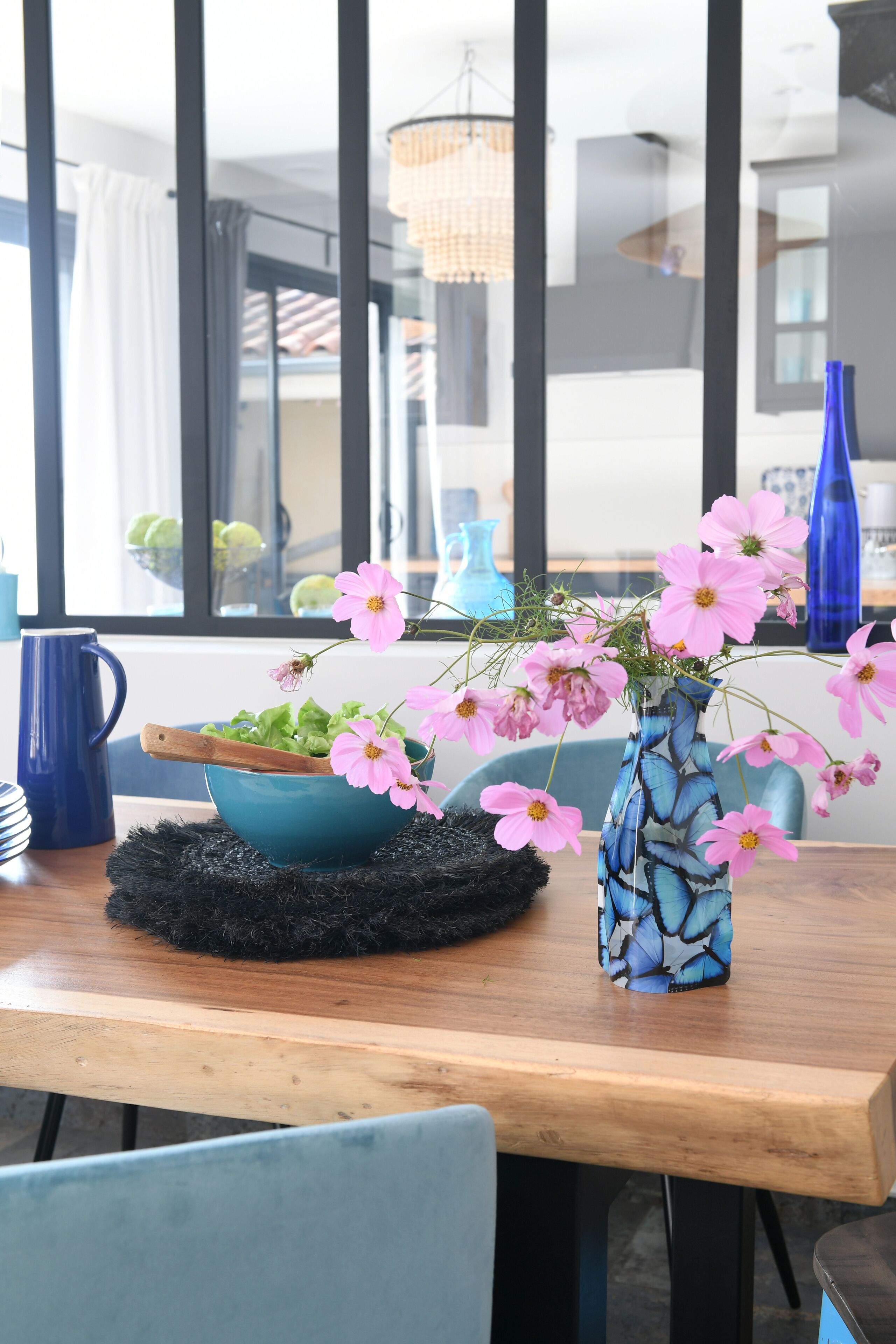 A dining table with a blue bowl and a vase with pink flowers, set on a black mat.