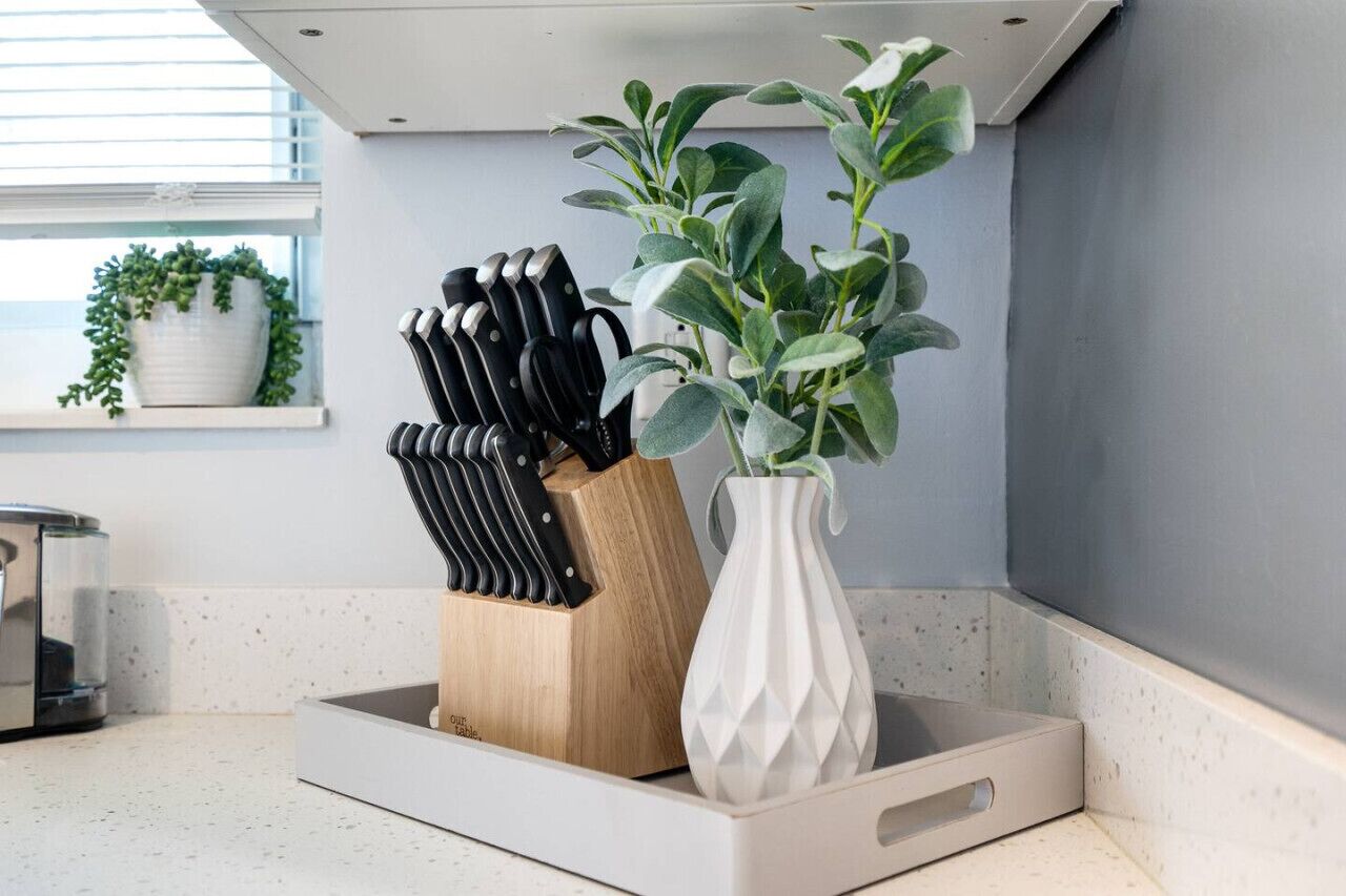 Een keukentafel met een messenblok, een witte vaas met groen loof en een plant in een potje op een raamkozijn.