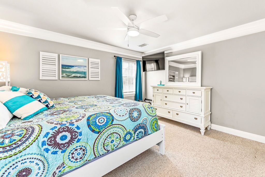 Une chambre à coucher comprenant un lit, une commode, un ventilateur de plafond et une fenêtre avec des rideaux.
