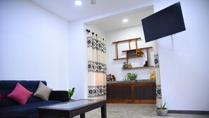 Ein Wohnzimmer mit einer Couch, einem Couchtisch, einem an der Wand befestigten Flachbildfernseher und einer Küchenzeile mit Arbeitsfläche und Regalen.