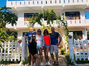 A group of people standing in front of a white building with balconies and a white picket fence.