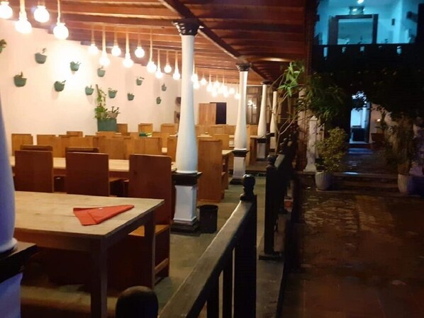 An indoor dining area with wooden tables and chairs, white pillars, and hanging light bulbs.