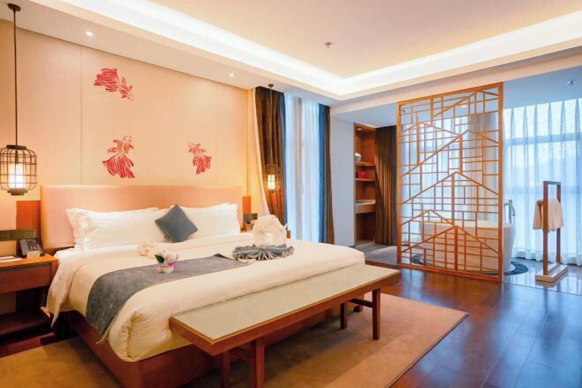 A hotel room with a large bed, bedside tables, a bench, a decorative screen, and a view of the outside.