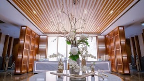 Um saguão com balcão de recepção, arranjo floral central e painéis de madeira nas paredes e teto.