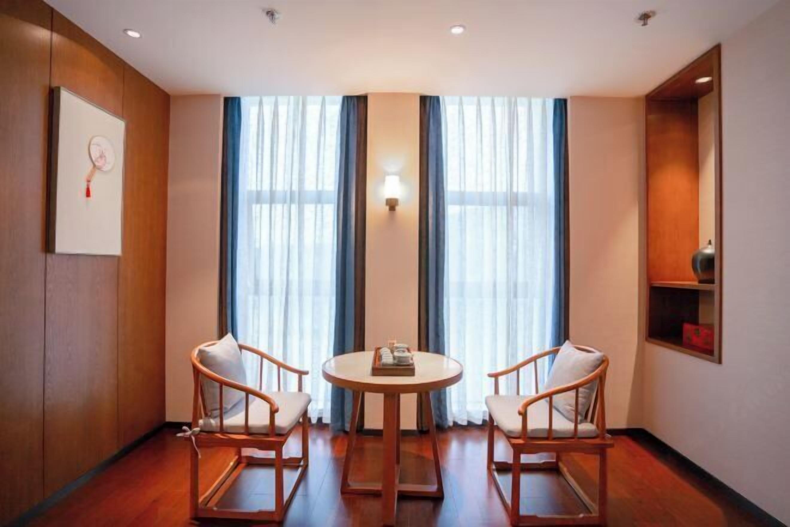 A room with a round table and two chairs, large windows with curtains, a wall-mounted light, and a shelf with a red object.