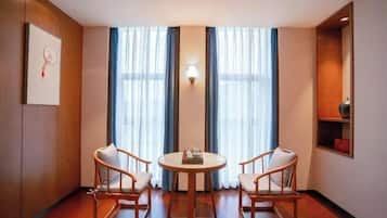 A room with a round table and two chairs, large windows with curtains, a wall-mounted light, and a shelf with a red object.