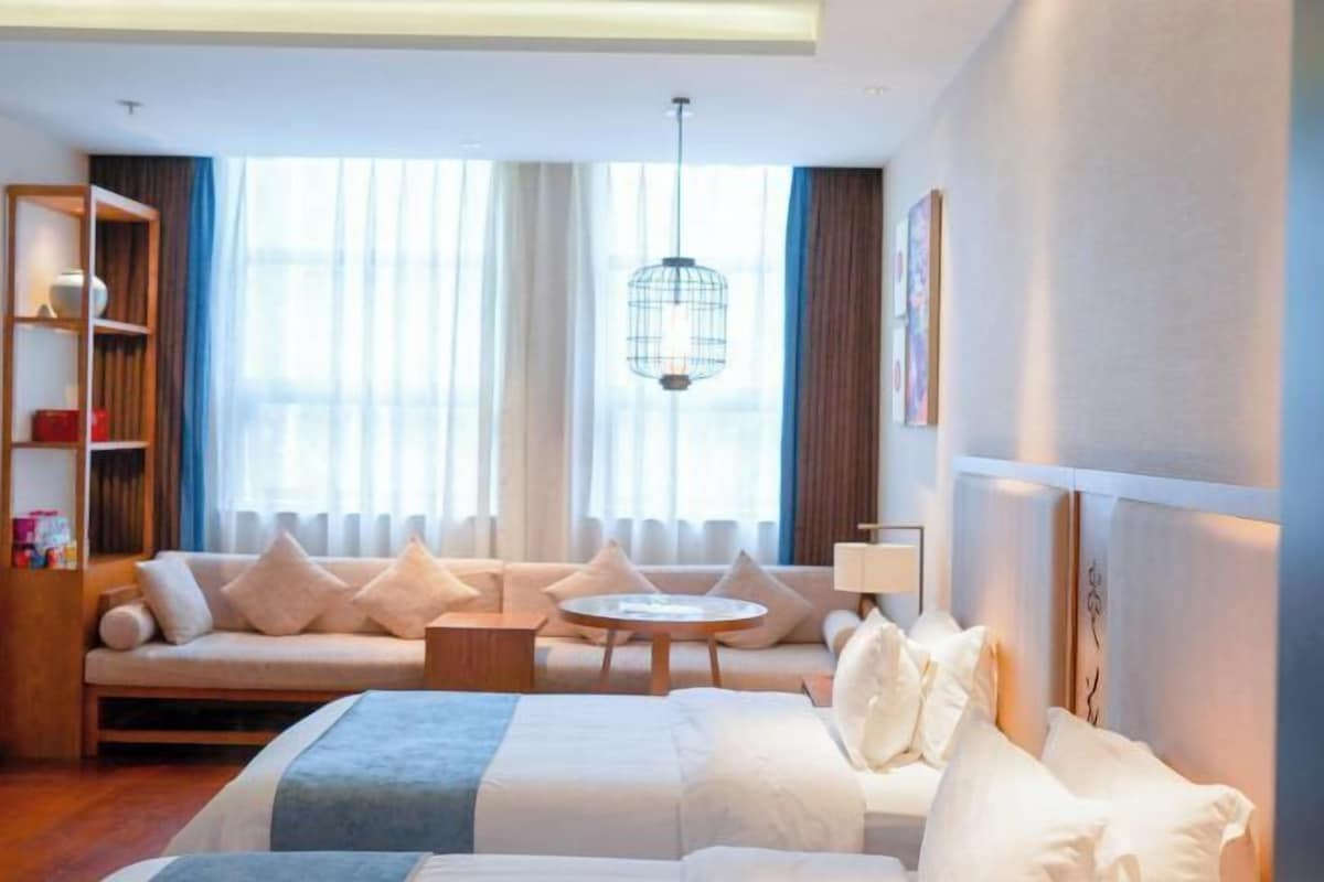 A hotel room with a bed, a sofa, a small table, a bookshelf, and a large window with curtains.