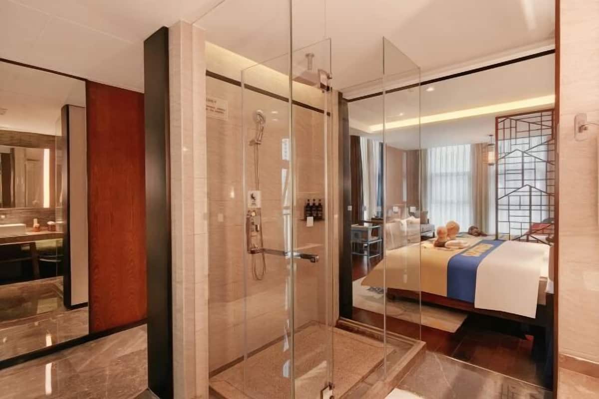 Una habitación de hotel moderna con ducha separada por vidrio, un espejo grande, una cama con colcha azul y blanca, y un panel de madera en la pared izquierda.