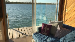 A wooden cabin with a sliding glass door leading to a wooden deck overlooking a lake. Inside, there is a blue sofa with cushions. - Sunset Point Houseboat, a gem of a place (Fresh Creek)