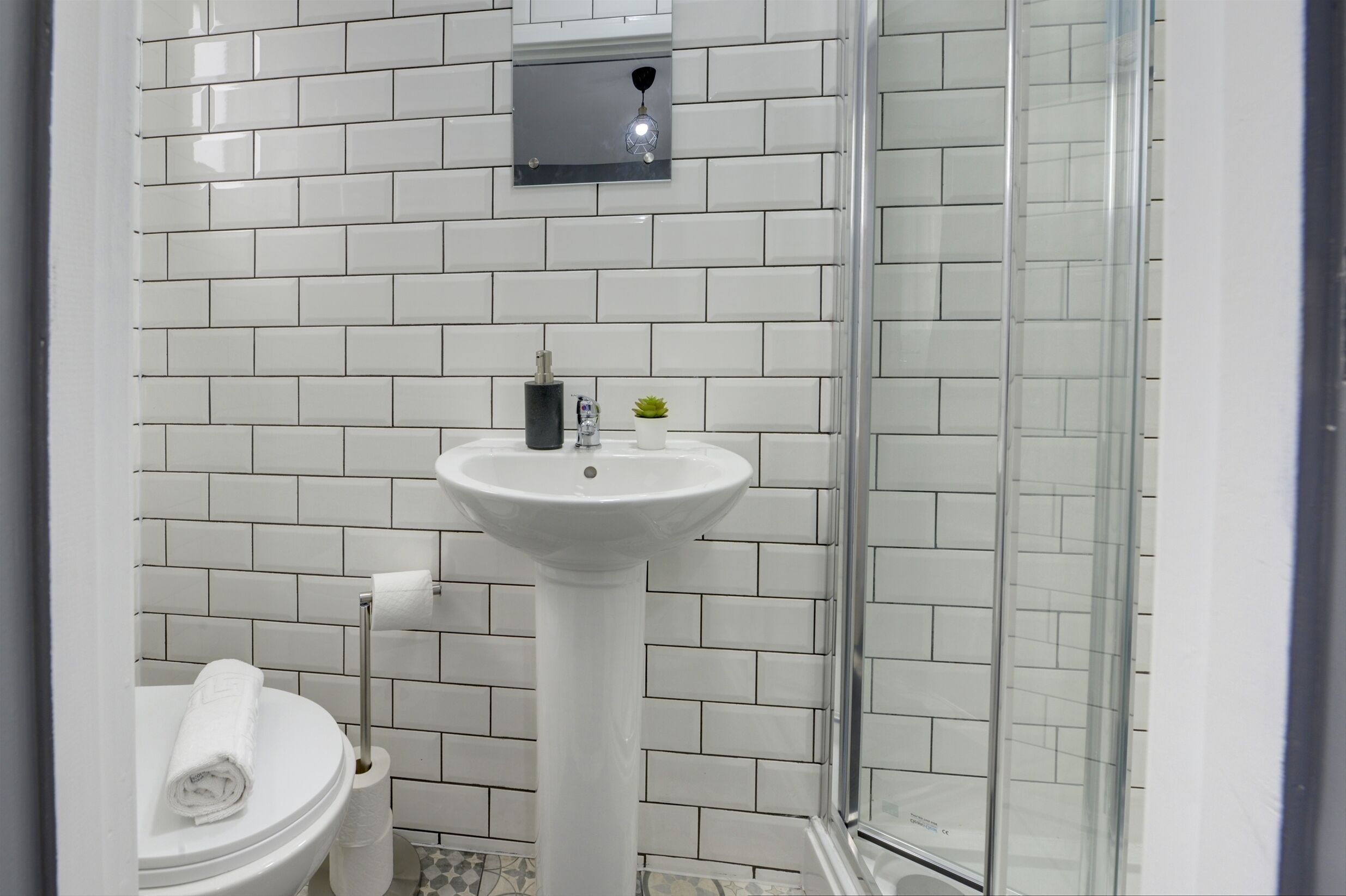 Un bagno con parete rivestita in piastrelle bianche, un lavabo su piedistallo, un water e una cabina doccia in vetro.