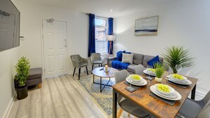 A modern living room with a sofa, chairs, a dining table set with plates and glasses, and a potted plant. - 56HRC 5 BedRoom House| Garden| Parking PFP (Coventry)