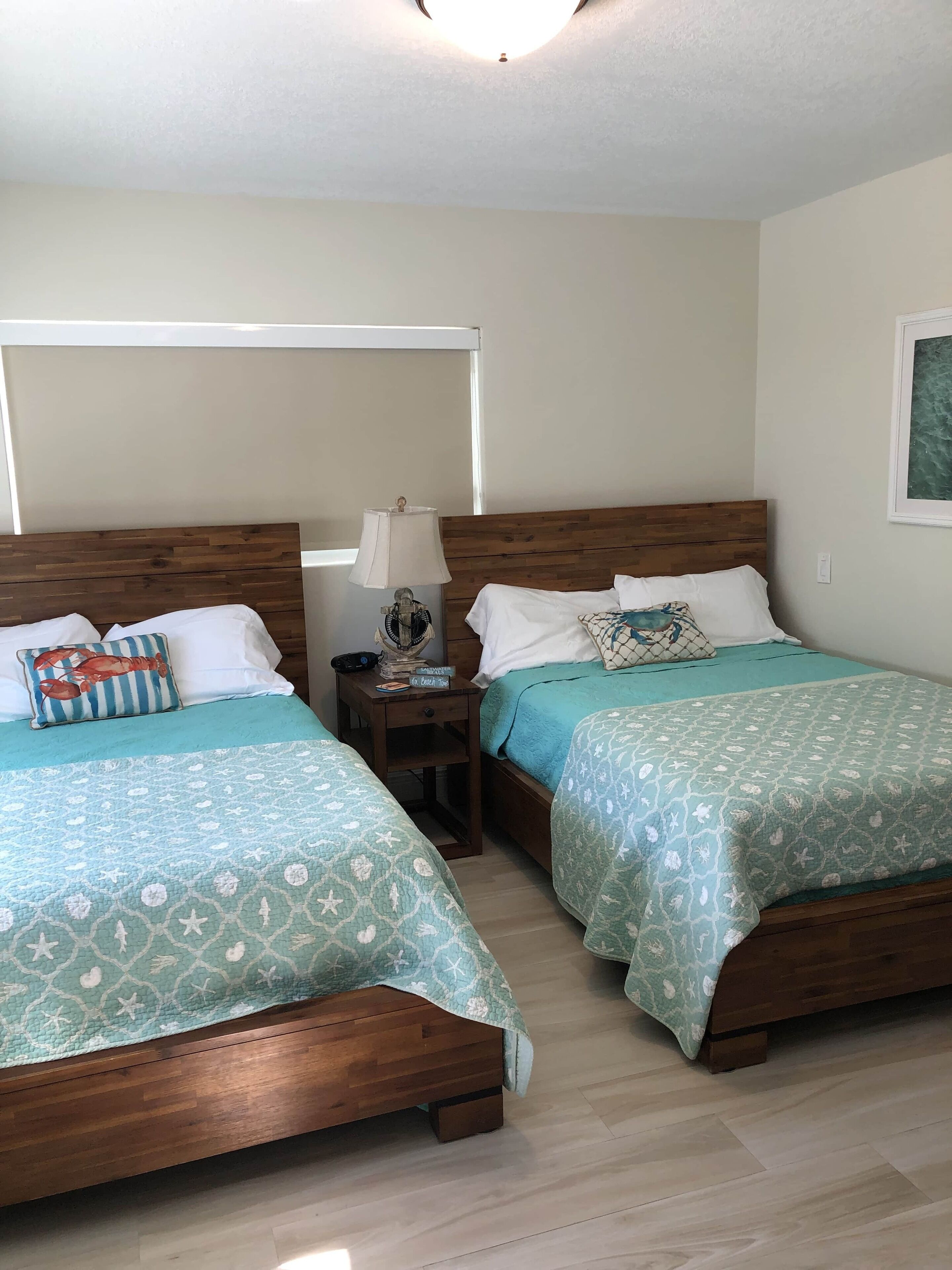 A hotel room with two wooden beds, each with a teal bedspread featuring a sea-themed pattern. There is a nightstand with a lamp and a small decorative item between the beds.