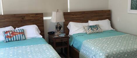 A hotel room with two wooden beds, each with a teal bedspread featuring a sea-themed pattern. There is a nightstand with a lamp and a small decorative item between the beds.