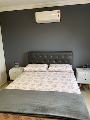 Un dormitorio con cabecera gris oscuro, colcha con estampado floral, manta gris y aire acondicionado blanco en la pared.