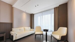 Una habitación de hotel moderna con un sofá, dos sillones, una mesita redonda y un ventanal con cortinas.