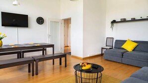 Un salon moderne avec un canapé gris, une table à manger avec des bancs, un téléviseur fixé au mur et une horloge.
