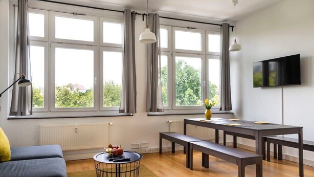 Un salon moderne doté de grandes fenêtres, d’un canapé, d’une table à manger avec des bancs, d’un téléviseur et d’une petite table ronde ornée d’un vase et de fruits.