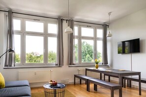 En moderne stue med store vinduer, en sofa, et spisebord med bænke, et fjernsyn og et lille rundt bord med en vase og frugt.